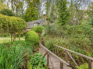 A wooden footbridge over a small stream with a garden and a brick house in the background at Saint Pancras Chapel in Washford near Williton