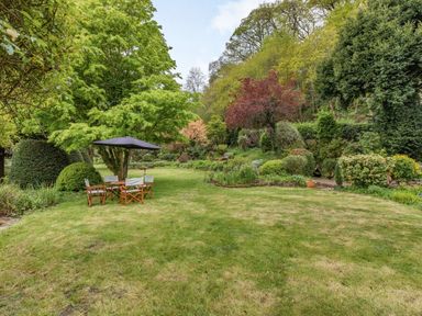 A garden with grass trees shrubs and a wooden table with chairs and a black umbrella at Saint Pancras Chapel in Washford near Williton