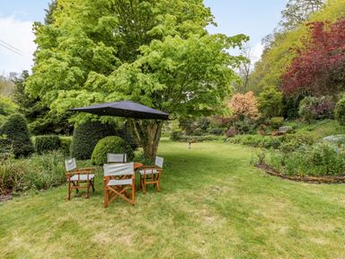 A garden with a table and chairs under an umbrella surrounded by trees and shrubs at Saint Pancras Chapel in Washford near Williton