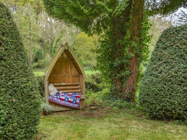 A wooden garden bench with a texas flag blanket and pillow between two large trimmed bushes and a tree at Saint Pancras Chapel in Washford near Williton