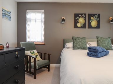 A bedroom with a bed with green pillows and folded towels on it a green armchair with a cushion beside a black chest of drawers and a window with blinds at Stonecroft in Praa Sands