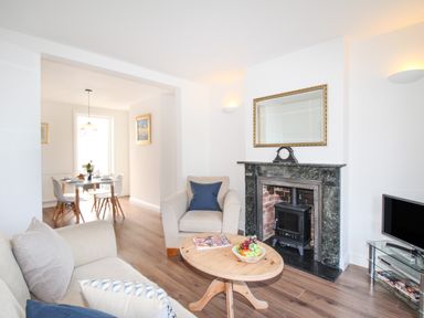 A living room with a wooden coffee table fruit bowl and magazines a beige armchair and sofa a fireplace with a clock and a dining table with chairs in the background at The Shambles in Fortuneswell