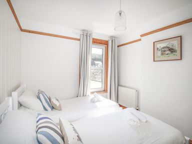 A small bedroom with two single beds white bedding striped pillows a window with curtains and a framed picture on the wall at The Shambles in Fortuneswell