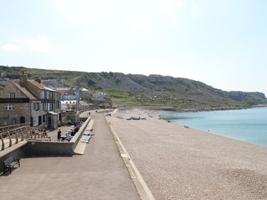 A pebble beach with people walking and sitting by the sea near a coastal town with buildings and hills in the background at The Shambles in Fortuneswell