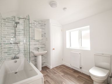 A bathroom with a bathtub shower, pedestal sink, toilet, window, radiator, and a door at The Shambles in Fortuneswell