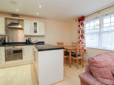 A kitchen with a black countertop island a wooden dining table with four chairs and a window with floral curtains at 1 Tregonwell Court in Minehead