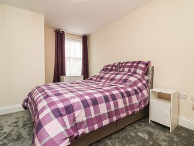 A bedroom with a double bed with checkered bedding beside a window with dark curtains and a white nightstand at 1 Tregonwell Court in Minehead