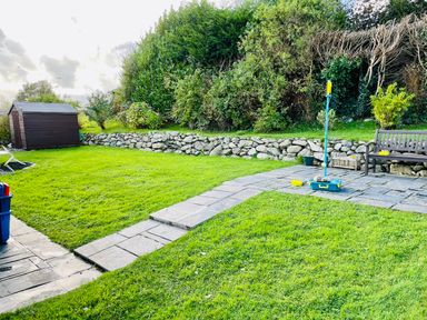 A backyard with a stone pathway a wooden shed green grass a bench and a stone wall at Yr Encil in Llanddaniel near Gaerwen