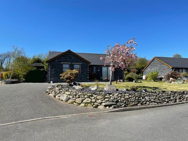 A stone house with a driveway and a garden with a flowering tree and a stone wall at Yr Encil in Llanddaniel near Gaerwen