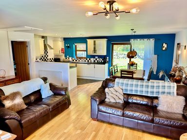 An open living room with two brown leather sofas a wooden dining table with chairs and a kitchen with white cabinets and blue walls at Yr Encil in Llanddaniel near Gaerwen