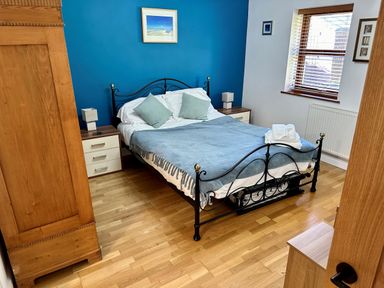 A bedroom with a black metal bed frame a blue wall two bedside tables with lamps a wooden wardrobe and a window with blinds at Yr Encil in Llanddaniel near Gaerwen