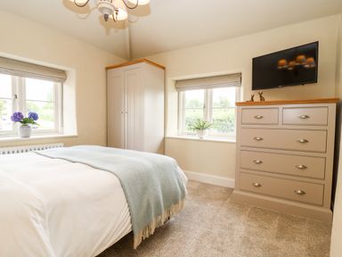 A bedroom with a bed covered with a blanket two windows plants on the windowsills a wardrobe a chest of drawers and a TV at 4 Lower Folley in Chipping Campden