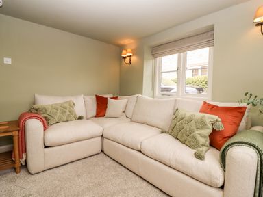 A corner sofa with cushions and throws in a living room with a window and wall lights at 4 Lower Folley in Chipping Campden