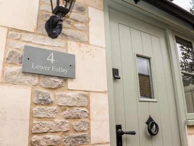 A green wooden front door with a small window and black handle next to a stone wall with a sign reading 4 Lower Folley and a black outdoor light fixture at 4 Lower Folley in Chipping Campden