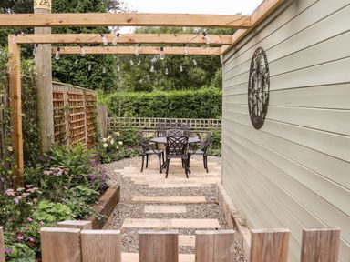 An outdoor garden area with a wooden pergola string lights metal table and chairs gravel ground and plants at 4 Lower Folley in Chipping Campden