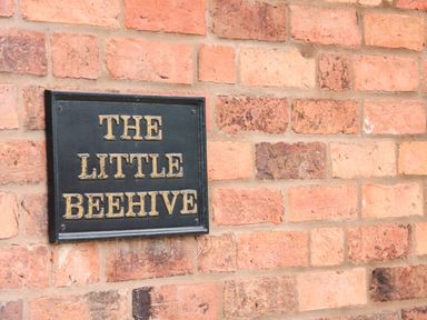 A black sign with gold lettering reading The Little Beehive mounted on a red brick wall at The Little Beehive in Abberley