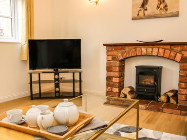 A living room with a television on a stand a brick fireplace with a stove and a glass coffee table with a tea set at The Little Beehive in Abberley