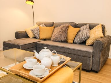 A gray sofa with yellow and patterned cushions next to a floor lamp and a glass coffee table with a wooden tray holding a white tea set at The Little Beehive in Abberley
