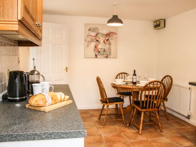 A kitchen with a countertop holding a loaf of bread and kitchen appliances and a dining table set with dishes and wine glasses at The Little Beehive in Abberley