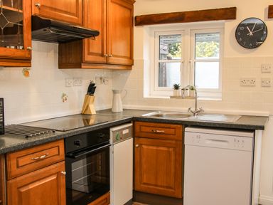 A kitchen with wooden cabinets a black stove a white dishwasher a sink under a window and a clock on the wall at The Little Beehive in Abberley