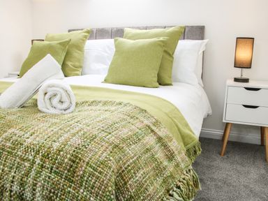 A bedroom with a bed with green pillows and a green blanket with two white rolled towels on it next to a white bedside table with a lamp at The Little Beehive in Abberley