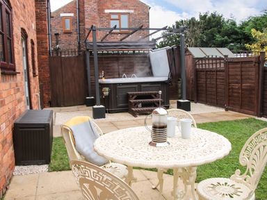An outdoor patio area with a white metal table and chairs a French press and two mugs on the table a hot tub with a pergola behind the table a brick wall and wooden fence enclosing the garden at The Little Beehive in Abberley
