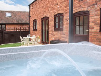 An outdoor patio area with a hot tub spraying water and a small white table with chairs next to a brick building at The Little Beehive in Abberley