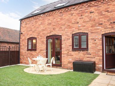 A backyard with a white metal table and chairs on a stone patio and a brick house with brown framed windows and doors at The Little Beehive in Abberley