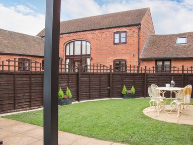 A backyard with green grass, a dark wooden fence, potted plants, and a white metal table with chairs at The Little Beehive in Abberley