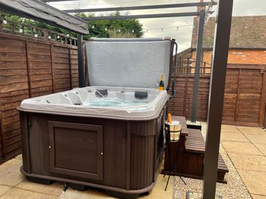 An outdoor hot tub with water jets under a pergola next to wooden steps and a bottle of champagne on a wooden fence at The Little Beehive in Abberley