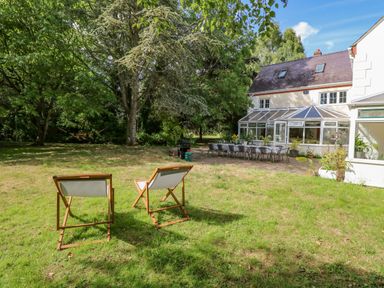 A garden with two deck chairs trees a long table and chairs and a white house with a conservatory at The Firs in Wendling near Dereham