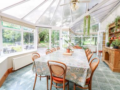 A dining area with a long table covered with a patterned tablecloth surrounded by wooden chairs in a glass conservatory with plants on a cabinet at The Firs in Wendling near Dereham