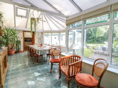 A dining area with a long table and several chairs surrounded by plants and large windows at The Firs in Wendling near Dereham