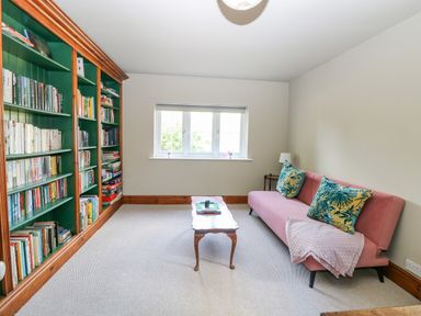 A room with a large bookshelf filled with books a pink sofa with cushions a wooden coffee table and a window at The Firs in Wendling near Dereham