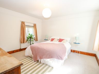 A bedroom with a double bed with pink and white bedding two bedside tables with lamps a striped rug and a window with curtains at The Firs in Wendling near Dereham