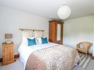 A bedroom with a double bed two teal pillows a wooden wardrobe a bedside table with a lamp and a wicker chair on a striped rug at The Firs in Wendling near Dereham