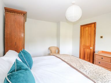 A bedroom with a bed with teal pillows a wooden wardrobe a wicker chair a wooden door and a wooden dresser at The Firs in Wendling near Dereham