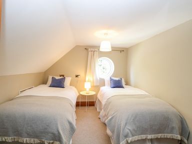 A bedroom with two single beds with grey blankets and blue pillows a round window and a small round table with a lamp at The Firs in Wendling near Dereham