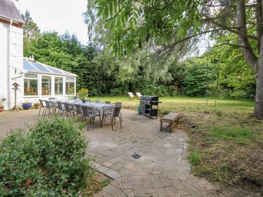 An outdoor patio with a long dining table and chairs a barbecue grill a wooden bench and a garden with trees and grass at The Firs in Wendling near Dereham
