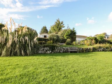 A garden with green grass a wooden bench and a house behind bushes at Tegfryn Bach in Llangoed near Beaumaris