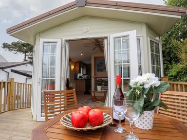 A wooden deck table with apples a wine bottle glasses and a flower pot outside of a building with open double doors at Tegfryn Bach in Llangoed near Beaumaris