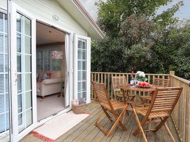 An outdoor wooden deck with a table and four chairs next to open glass doors showing a living room with a sofa and lamp at Tegfryn Bach in Llangoed near Beaumaris