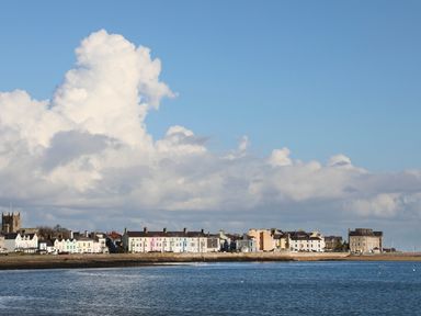 A coastal town with a row of buildings along the shore and a church tower under a cloudy sky at Tegfryn Bach in Llangoed near Beaumaris
