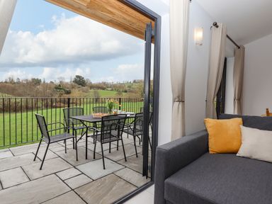 A patio dining table with six chairs and flowers overlooking green fields next to a room with a gray sofa and curtains at Woodpecker Lodge in Butterleigh
