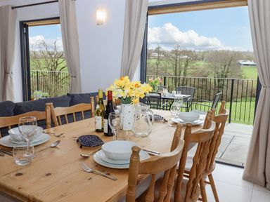 A dining area with a wooden table set with plates glasses and flowers next to large windows overlooking a balcony and green landscape at Woodpecker Lodge in Butterleigh