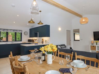 A kitchen and dining area with a wooden table set with dishes and yellow flowers at Woodpecker Lodge in Butterleigh