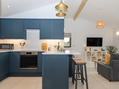 A kitchen with a blue island and bar stools next to a living area with a grey sofa and television at Woodpecker Lodge in Butterleigh
