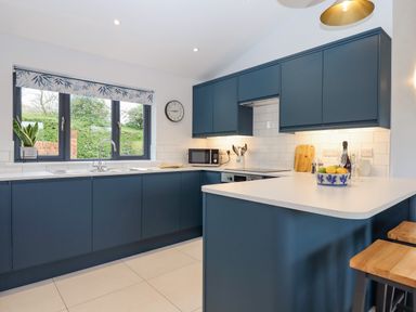 A kitchen with blue cabinets white countertops a window with a plant a clock and stools at Woodpecker Lodge in Butterleigh