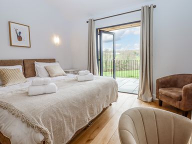 A bedroom with a double bed featuring pillows and towels next to an open door with curtains and a view outside at Woodpecker Lodge in Butterleigh