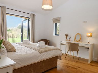 A bedroom with a bed and white towels on it a desk with a chair and a lamp and an open door leading outside at Woodpecker Lodge in Butterleigh
